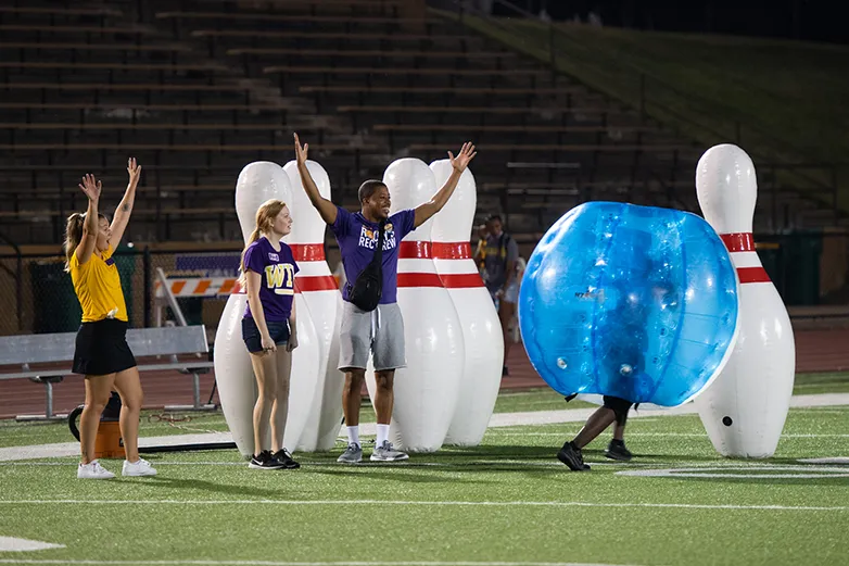 three students, two with their arms up celebrating, in front of giant bowling pins that appear to be part of a game being played on Hanson Field